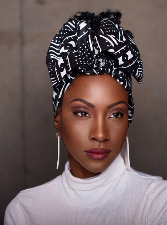 A stylish woman wearing a black and white patterned wire headwrap tied into a large front bow. She has on long textured statement earrings, and a white turtleneck top. Her makeup is polished with deep red lipstick, creating a modern, elegant look.